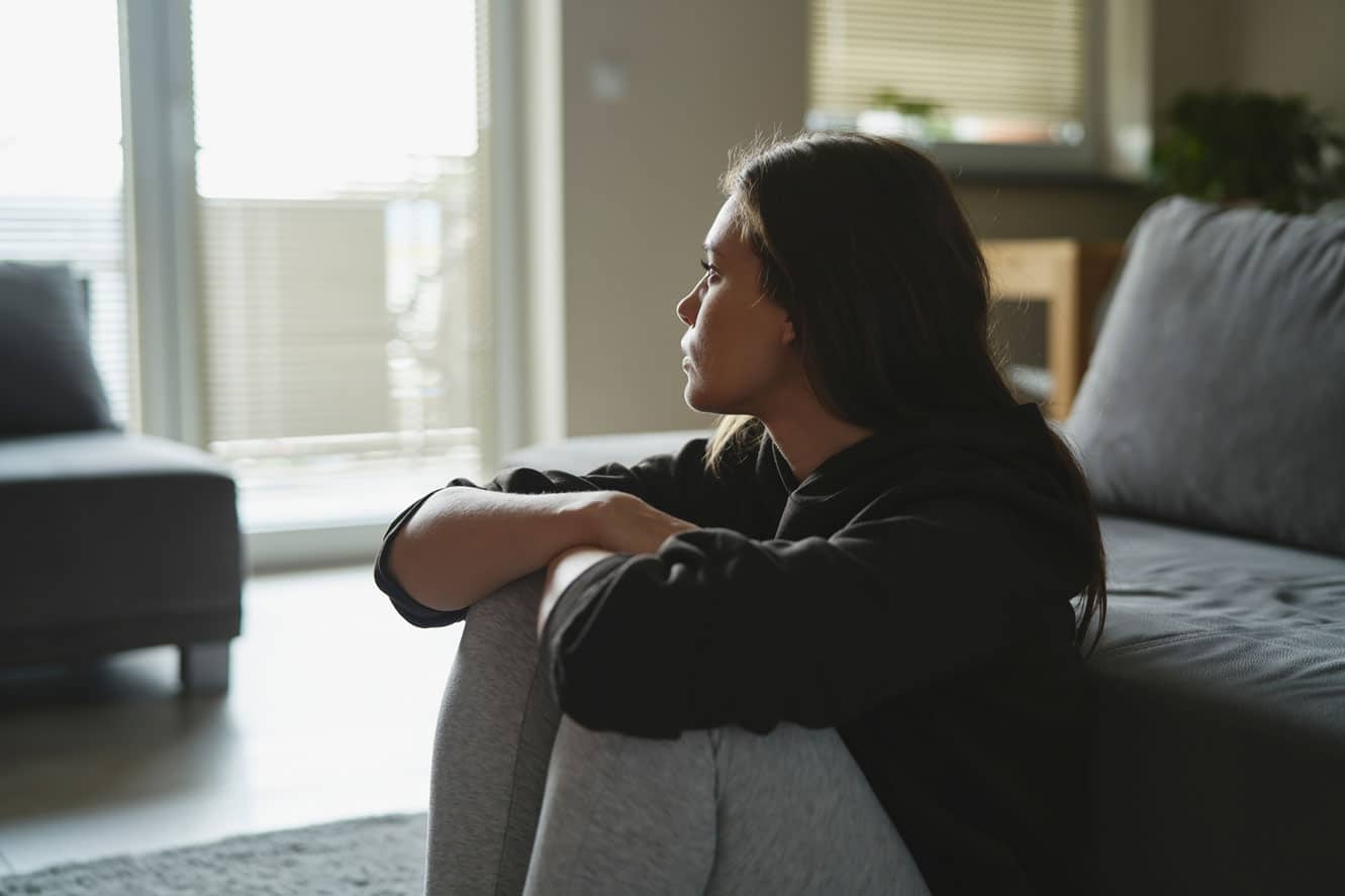Woman sitting on the floor against the couch, knees drawn up to her chest.