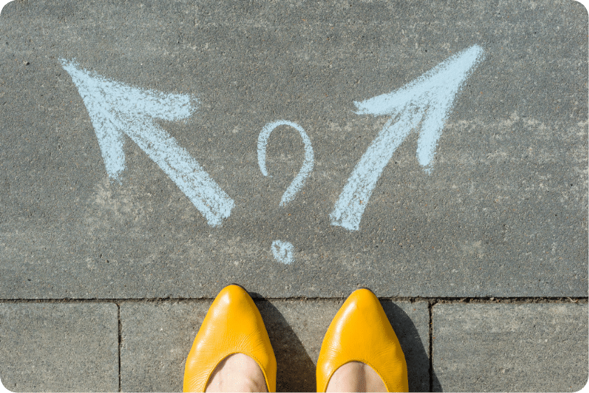 Yellow shoes standing on pavement, in the middle of two chalk drawings of arrows pointing into two different directions, with a question mark drawn in the middle.