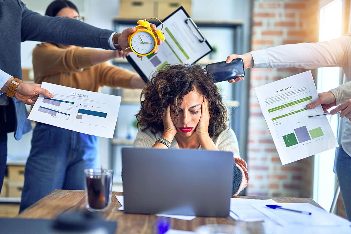 Woman sitting in front of her laptop looking stressed with hands holding documents pointing at her from all directions.